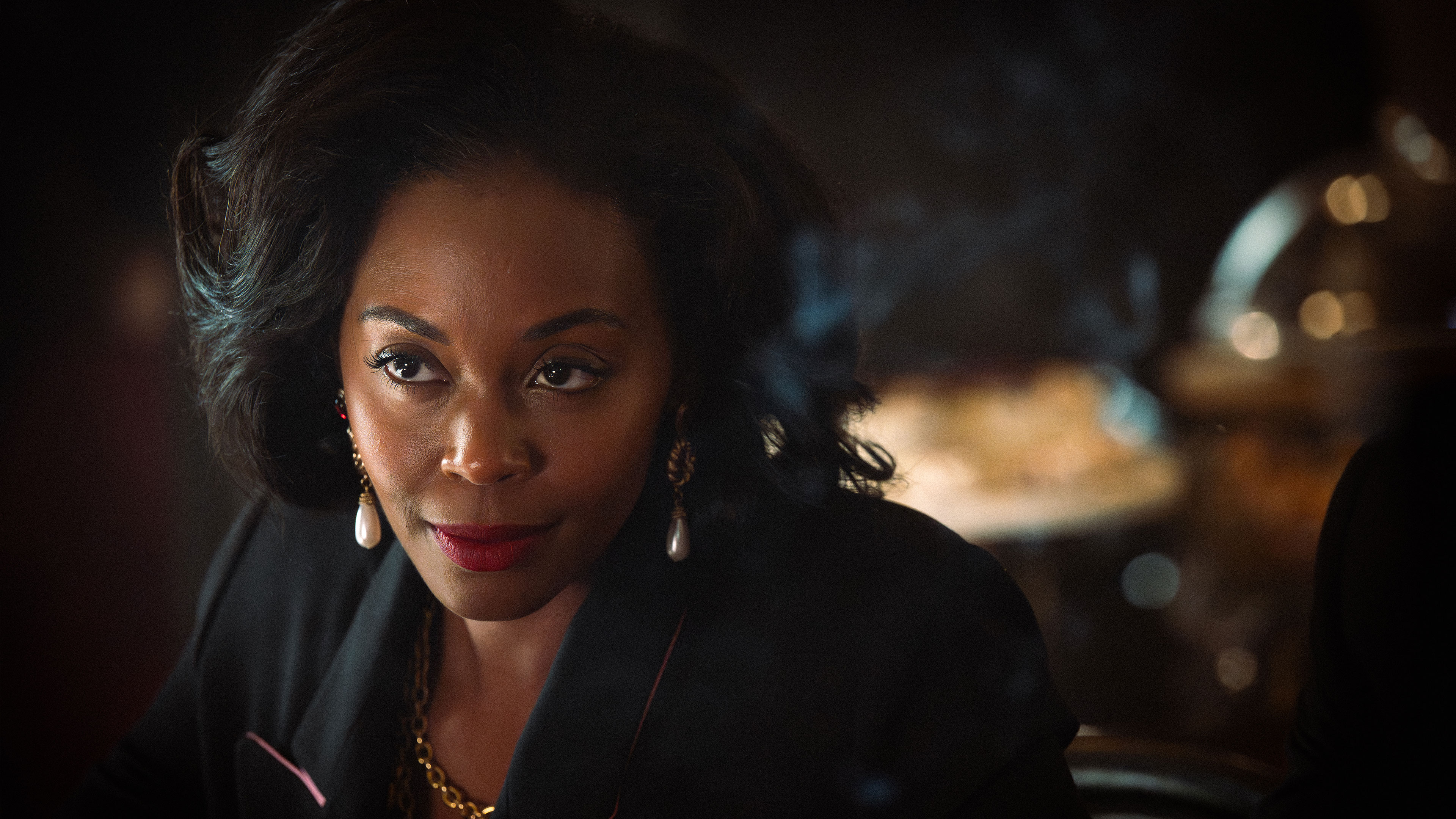 Cameron Cook (Nafessa Williams) in close-up wearing gold jewellery, lit in a dim interior setting in Rivals season 2.
