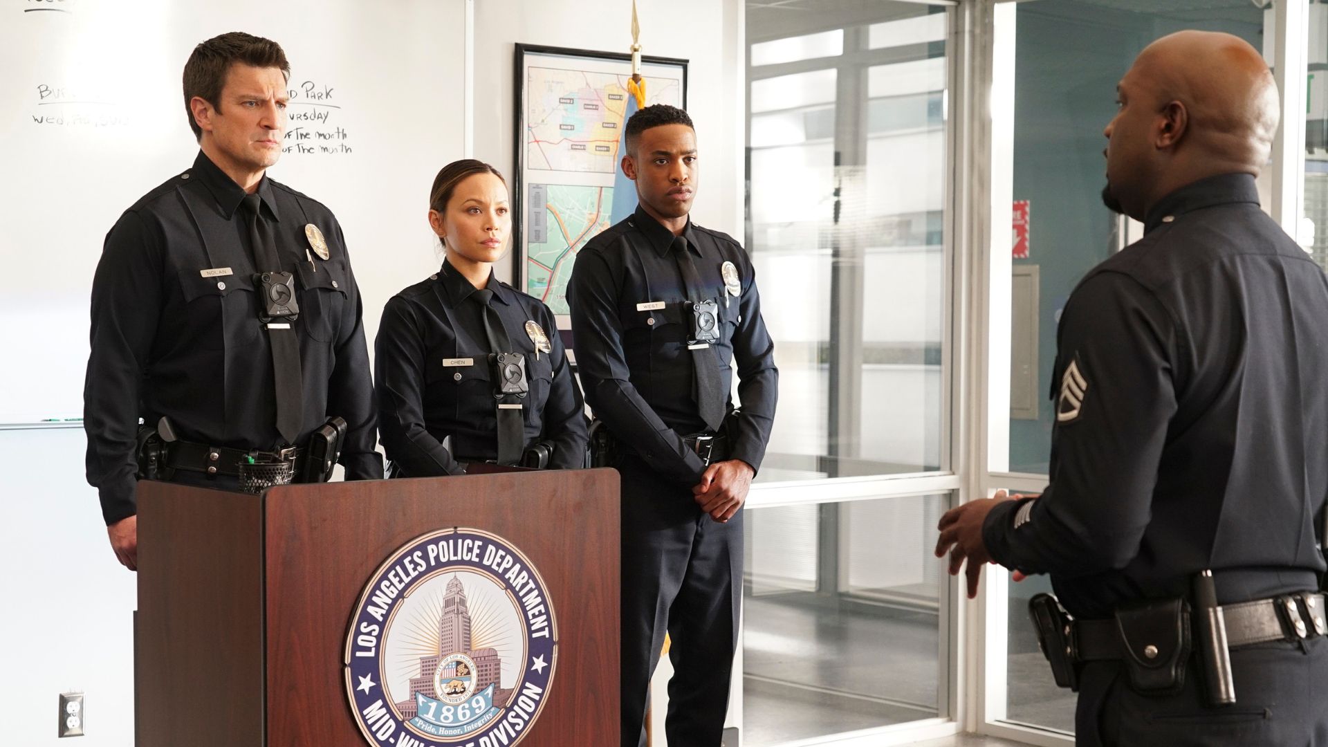 Officers Nolan, Chen, and West stand at attention behind a podium as their sergeant addresses them in a scene from The Rookie.