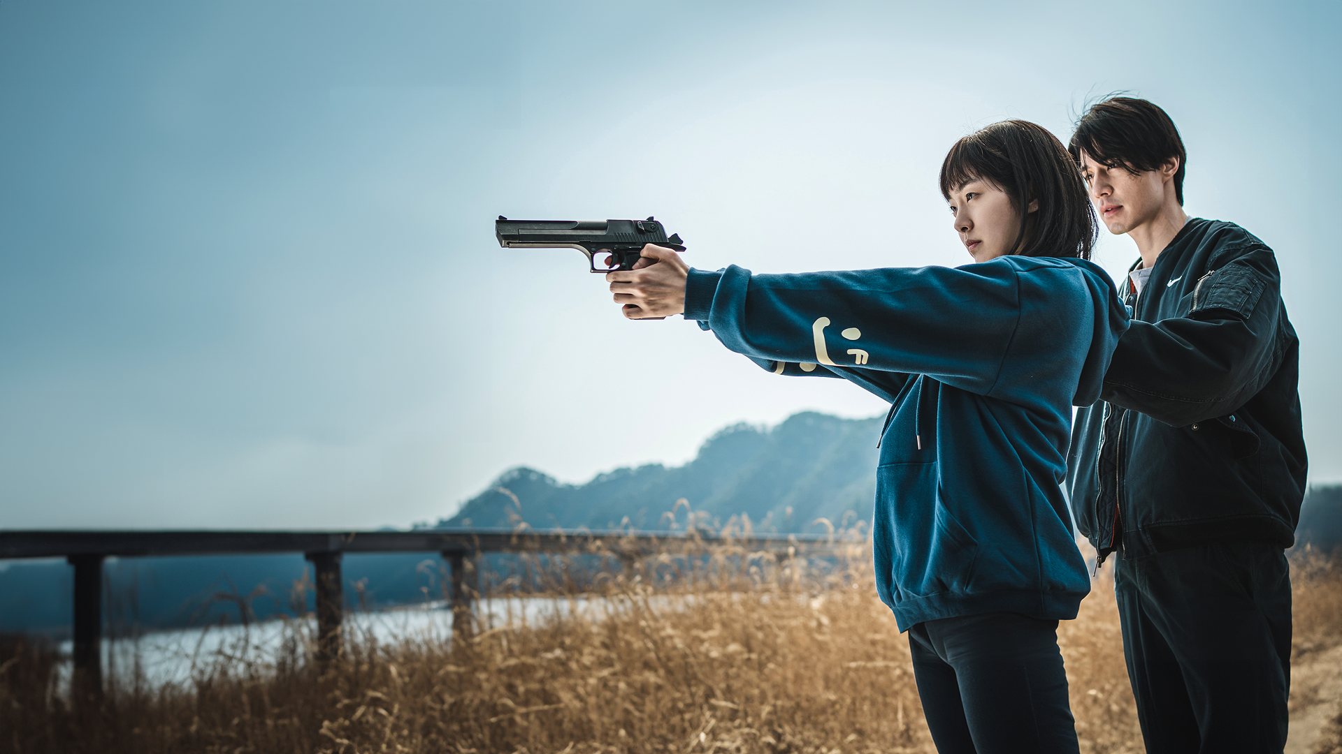 A dramatic promotional image from "A Shop for Killers" showing a young woman in a blue hoodie holding a large pistol with both hands, aiming it forward with focus, while a man in a black jacket stands closely behind her, guiding her posture against an outdoor background of dry grass, hills, and an overcast sky.