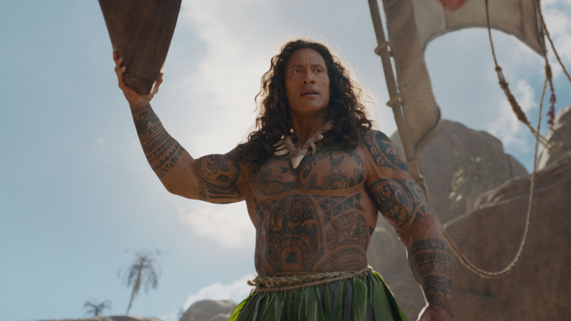 Dwayne Johnson as a live-action Maui stands shirtless with a serious expression, displaying his extensive tribal tattoos and wearing a leaf skirt under a bright, partly cloudy sky.