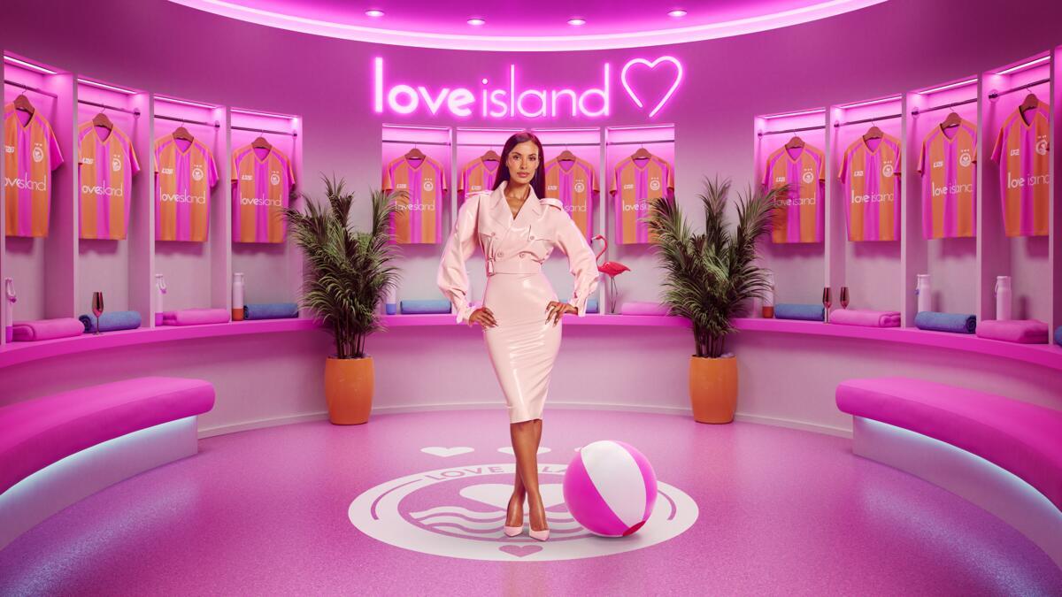 Maya Jama stands in the pink dressing room set for Love Island UK Season 13, surrounded by team jerseys and neon Love Island branding.