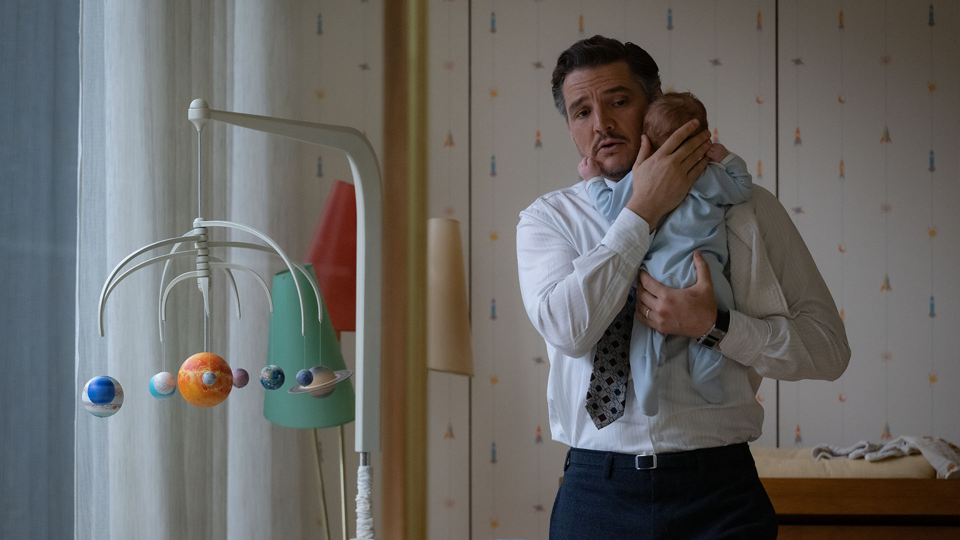 A cinematic scene from The Fantastic Four: First Steps shows Reed Richards (Pedro Pascal) looking concerned while gently cradling a baby in his arms, with a colorful solar system baby mobile hanging in the foreground.